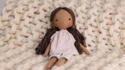 A hand-sewn doll with tan skin and brown hair in a light pink shirt and grey pants sitting on a blanket, all made in Katia Ferris's Sew a 14-Inch Rag Doll class on Creativebug.