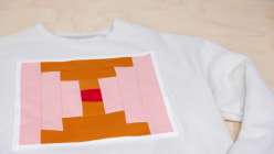 A cream colored sweatshirt with an ochre and pink quilt block stitched to the front, made in Faith Hale's Make a Quilt Block Sweatshirt class on Creativebug.