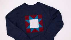 A navy blue sweatshirt with a blue, burgundy, and white quilt block stitched to the front, made in Faith Hale's Make a Quilt Block Sweatshirt class on Creativebug.